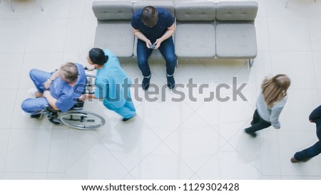 High Angle Shot in the Hospital Lobby, Young Man Waits for Results while Sitting and Using Mobile Phone, Doctors, Nurses and Patients Walk Past Him. Clean, New Hospital. Royalty-Free Stock Photo #1129302428