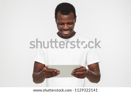 Studio shot of young African American man pictured isolated on gray background wearing blank white t shirt looking at screen of tablet he is holding, smiling happily