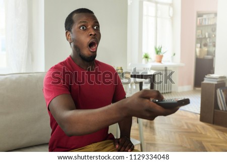 Indoor picture of young handsome dark-skinned man spending leisure time at home switching TV programs looking deeply astonished with some shocking news, showing strong emotions of surprise on his face