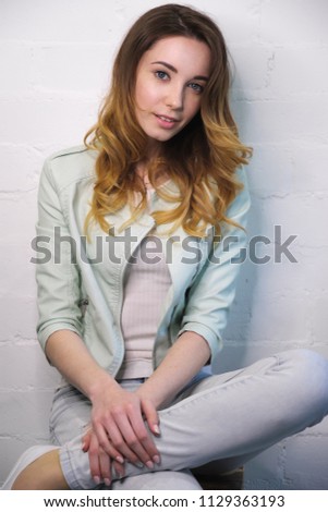 Portrait of a seated girl with a tossed foot on her leg against a white wall looking at the camera. Vertical photo