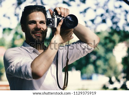 The photographer is working. Guy with smiling face takes picture outside. Photographer holds retro camera. Man with beard holds photocamera on green nature background.