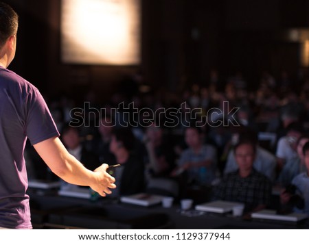 Presenter Speaking to Audience People in Conference Hall Auditorium. Presentation Stage. Blurred De-focused Unidentifiable Audience and Presenter. Technology. Casual Attire Presenter. Royalty-Free Stock Photo #1129377944
