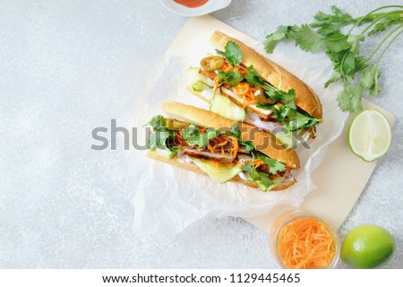 Classical banh-mi sandwich with sliced grilled pork tenderloin, shredded carrots and peeled cucumbers, jalapeno peppers and cilantro on white textured background. Top view, copy space Royalty-Free Stock Photo #1129445465