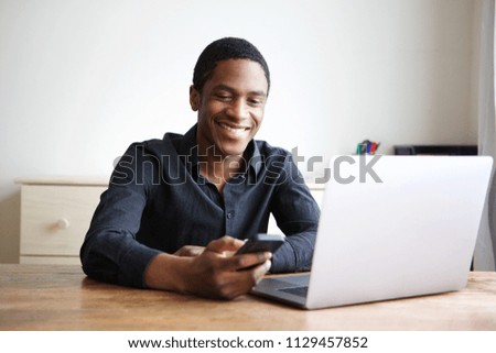 Portrait of happy young businessman sitting at desk with computer and sending text on mobile phone