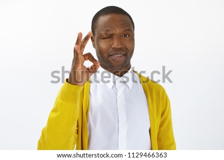 Don't worry, everything will be fine. Handsome young Afro American guy with bristle blinking at camera and making ok gesture as sign of approval, like and positive attitude, expressing good emotions