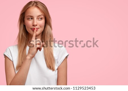 Indoor shot of beautiful female looks mysteriously aside, has intriguing look, asks to be quiet, dressed in casual clothes, stands against pink background with blank copy space for your advertisement Royalty-Free Stock Photo #1129523453