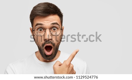 Close up shot of emotive shocked bearded young male opens mouth widely, stare at camera, points upwards aside at blank space, says: Wow, it`s something unbelievable! People and emotions concept Royalty-Free Stock Photo #1129524776