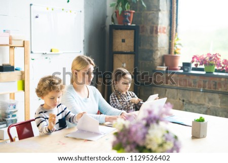 Sunlit portrait of young woman working at laptop with two kids painting beside her at table in modern apartment, copy space