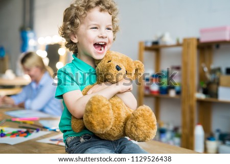 Warm toned portrait of happy curly haired kid laughing cheerfully and hugging teddy bear toy, copy space Royalty-Free Stock Photo #1129526438