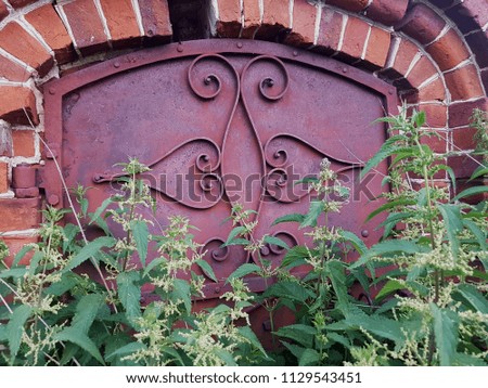 An old, vintage building made of red brick, a red door in patterns. Grass nettle in the foreground.