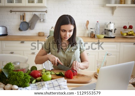 Charming young brunette woman in casual shirt recording video for her culinary blog, sitting at kitchen table in front of open laptop and cutting red bell pepper while making vegetable salad