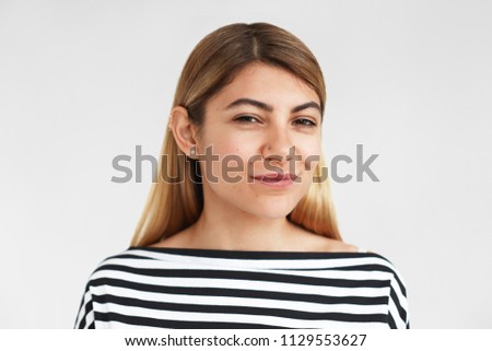 Picture of charming fashionable blonde hipster girl wearing striped top and facial piercing looking at camera with mysterious playful smile, enjoying leisure time, having pleased carefree look