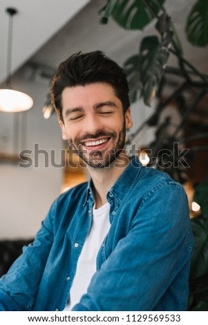 Portrait of bearded hipster man with toothy smile and fashion hairstyle sitting in loft modern coffee shop. Satisfied customer wearing casual clothes posing for pictures, good mood, feeling happy. 