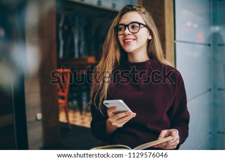 Happy hipster girl wear in spectacles for vision correction sitting at cafeteria with loft interior and holding modern smartphone and waiting for call, concept of technology and communication Royalty-Free Stock Photo #1129576046