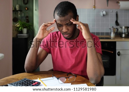 Indoor shot of young African American man pressing fingers to temples feeling pain in his head trying to figure out accounting details of his bills, looking helpless and uneasy, not knowing what to do Royalty-Free Stock Photo #1129581980