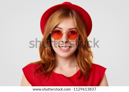 Close up shot of pretty Caucasian young female with gentle smile, looks happily at camera, wears red shades and hat, rejoices recieving compliment, isolated on white background. Fashion concept