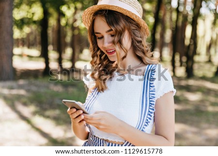 Glamorous curly girl in trendy clothes looking at phone screen. Outdoor shot of fascinating female model in hat texting message after photoshoot in park.