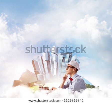 Young man writer in hat and eyeglasses using typing machine while sitting at the table with floating city and cloudy skyscape on background.