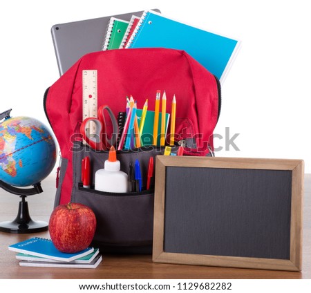 Backpack with an assortment of school supplies with a blank chalkboard on a wooden desktop and white backkground