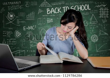 Picture of tired female college student reading a book and sitting with scribbles in the chalkboard