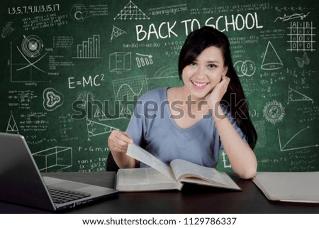 Picture of female college student smiling at the camera while reading a book and sitting with scribble in the chalkboard 