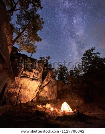 Fantastic landscape at summer night. Brightly burning small campfire in canyon amid huge steep rock formation under clear dark starry sky. Tourism, safety, climbing, hiking and traveling concept.