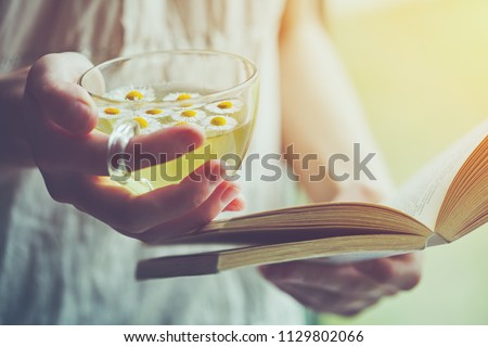 young woman holding cup of chamomile tea and reading open book Royalty-Free Stock Photo #1129802066