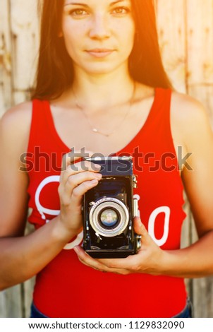 young beautiful brunette girl in red shirt and jeans shorts posing with a camera on the rustic wooden background. making photos, taking pictures, woman, photographer,vintage camera, outdoor portrait