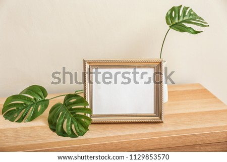 Photo frame with vase and green leaves on table near light wall
