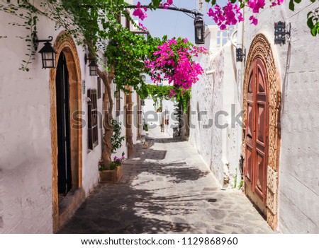 Narrow street in Lindos town on Rhodes island, Dodecanese, Greece. Beautiful scenic old ancient white houses with flowers. Famous tourist destination in South Europe Royalty-Free Stock Photo #1129868960