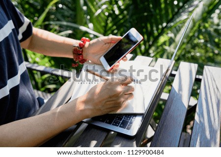 Man's hand holding a pencil and writing in notebook on workspace table with computer and mobile phone