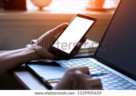 Mock up businessman hand holding blank screen mobile smart phone and browsing internet on laptop computer in office.