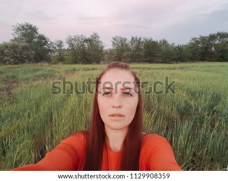young beautiful woman in red dress and red hair, takes a selfie on the phone in the green wheat field in the evening at sunset in summer. the nature of digital technology.