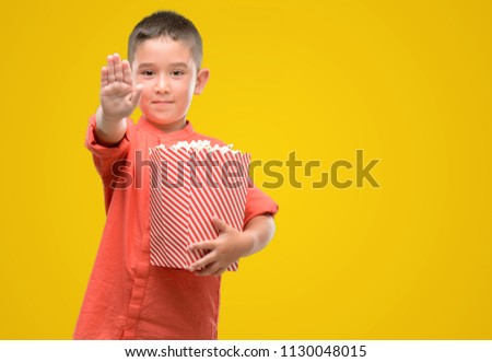 Dark haired little child eating popcorn with open hand doing stop sign with serious and confident expression, defense gesture