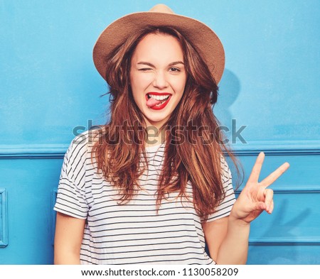Young stylish girl model in casual summer clothes  and brown hat with red lips, posing near blue wall. Winking and showing peace sign