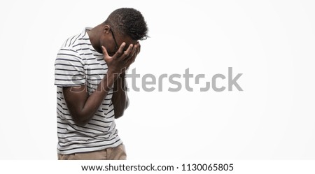Young african american man wearing glasses and navy t-shirt with sad expression covering face with hands while crying. Depression concept.