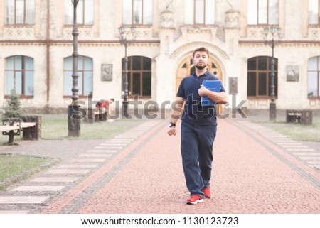 Happy man student goes with books in his hands on campus background. Portrait of a full-grown,funny man with a beard and books in his hands walking along the alley on the background of the university