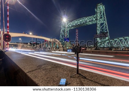 Camera on stabilizer shooting a time lapse of traffic light trails at night on a bridge with the Old monumental railroad bridge De Hef,  Rotterdam on the background
 
