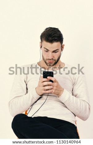 Guy with beard holds MP3 player and wears earphones on white background. Macho with headphones and cell phone listens to music. Man with surprised face looks at phone. Music and technology concept