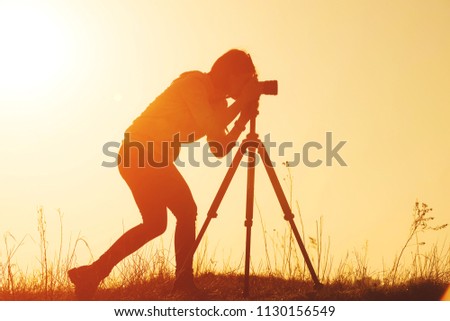 Silhouette of woman photographer shooting landscape with camera on tripod at sunset time in the field