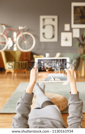 Tablet in the hands of a young woman lying down with her legs on the wooden floor and green carpet in a multifunctional room interior with vintage furniture in the blurred background