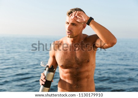 Picture of handsome mature sportsman standing with bottle of water drinking on the beach.