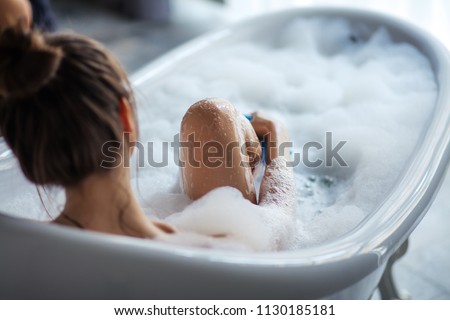 female massaging her legs with sponge in the tub. back view shot