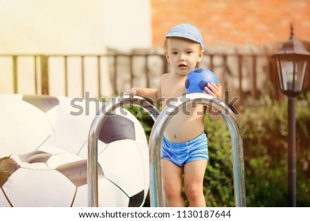 Child in swimming (bathing) trunks stands near swimming pool with a ball in hot summer day. Soccer ball decorated bean bag chair in background. Toned image.