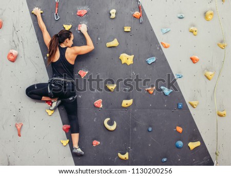 Female fitness professional climber training at bouldering gym. Muscular woman with athletic body dressed in black, climbing on artificial colourful rock wall. Active lifestyle and bouldering concept. Royalty-Free Stock Photo #1130200256