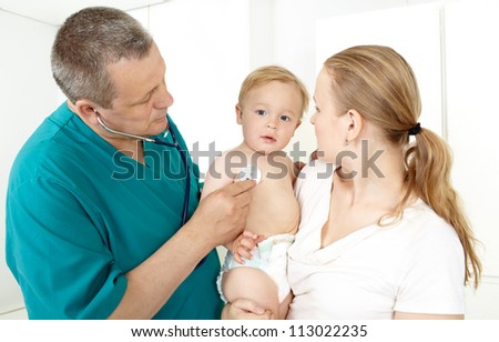 Doctor, middle aged, in a green smock, is checking a baby with a phonendoscope in a light medical study. The baby is 16 months old. He is sitting on his mum's laps. Caucasian, white.