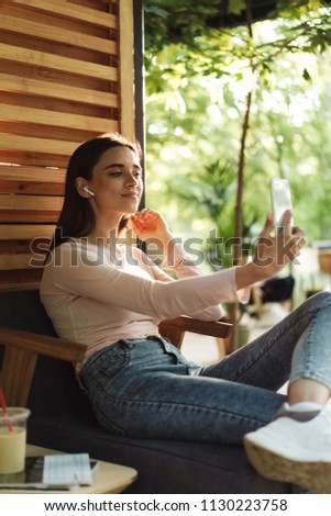 Pretty young girl in wireless earphones taking a selfie while sitting indoors at a cafe