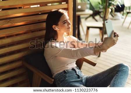Smiling young girl in wireless earphones taking a selfie while sitting indoors at a cafe