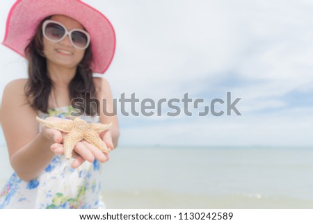 Beautiful woman wearing hat beach and sunglasses and holding starfish on hands over sandy beach, green sea and blue sky background for summer holiday and vacation concept.