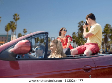 leisure, road trip, travel, summer holidays and people concept - happy friends driving in convertible car and taking picture by film camera over venice beach background in california